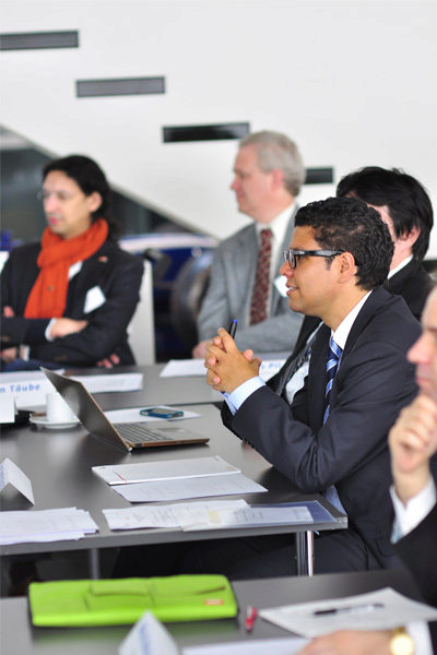 A small group of people in a business meeting, seated at a table with laptops and documents. One person is attentively listening, hands clasped.