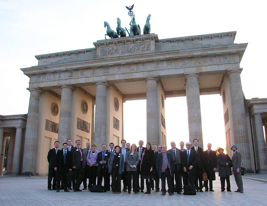 A large group of people in formal attire pose in front of the Brandenburg Gate in Berlin, Germany.